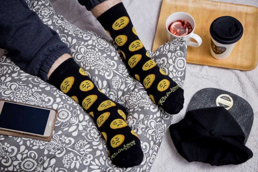 A person in Emoji Socks, sitting on the bed, drinking tea